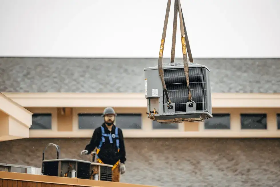 A packaged rooftop unit hanging from crane straps over a commercial roof